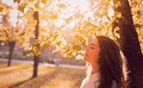 Rechts steht eine Frau mit geschlossenen Augen in der Herbstsonne, im Hintergrund herbstliche Bäume