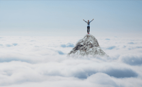 Eine Person steht mit erhobenen Armen auf einem Berggipfel, der aus einer Wolkendecke ragt.