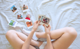 Eine Person sitzt im Schneidersitz auf einem Bett und betrachtet mehrere Sofortbilder mit Urlaubsmotiven.
