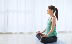 Eine Frau sitzt in Yogahaltung (Lotussitz oder ähnliches) auf einer Matte und meditiert.