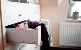 Nahaufnahme einer halb geöffneten Schublade einer weißen Kommode, aus der bunte und dunkle Kleidung herausquillt. Im Hintergrund sind ein Fenster, eine Heizung und eine hellrosa Wand zu erkennen.