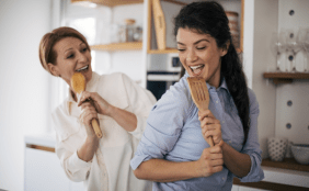 Zwei Frauen (eine mittleren Alters, eine jüngere) singen und lachen ausgelassen in einer modernen Küche, wobei sie hölzerne Küchenutensilien wie Löffel und Pfannenwender als Mikrofone benutzen.