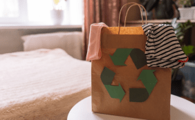 Eine braune Papiertüte mit dem grünen Recyclingsymbol, gefüllt mit Kleidung, steht auf einem Tisch. Im Hintergrund ist ein leicht verschwommenes Schlafzimmer mit einem hellen Bett zu sehen.