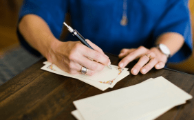 Person in blauem Oberteil sitzt an einem dunken Tisch und schreibt auf ein schönes Briefpapier