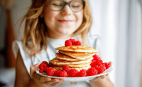Nahaufnahme eines lächelnden Mädchens mit Brille, das einen Teller mit einem Stapel Pfannkuchen hält, die reichlich mit frischen Himbeeren garniert sind.