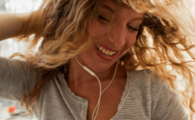 Eine junge Frau mit lockigem Haar lacht und bewegt sich freudig, während sie kabelgebundene Kopfhörer trägt.