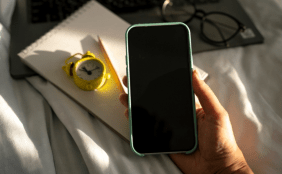 Eine Hand hält ein Smartphone mit schwarzem Bildschirm. Im Hintergrund liegen ein Notizblock, ein gelber Wecker und eine Brille auf einem hellen Laken.