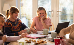 Mehrere Frauen an einem Tisch, vor ihnen verschiedene Projekte und Gegenstände. sie lachen und wollen gemeinsam Dinge angehen