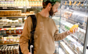 Mann im beigen Pulli und Rucksack und Bart steht im Supermarkt am Kühlregal