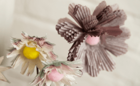 Nahaufnahme von kunstvollen, selbstgebastelten Papierblumen. Die Blütenblätter bestehen aus pastellfarbenen Papier-Muffinförmchen mit weichen Pompons als Blütenmitte in Gelb und Rosa.