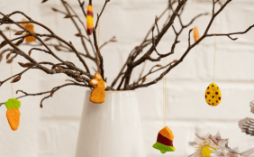 Ein weißer Krug mit kahlen Zweigen, an denen handgemachte Anhänger aus Salzteig oder Ton hängen. Zu sehen sind bunte Motive wie ein Hase, ein Ei mit Punkten und eine Karotte.