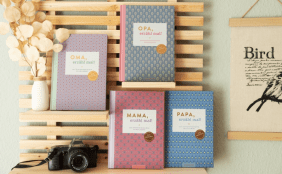 Vier verschiedene Erinnerungsalben von Elma van Vliet („Oma, erzähl mal!“, „Opa, erzähl mal!“, „Mama, erzähl mal!“, „Papa, erzähl mal!“) sind dekorativ in einem hellen Holzregal aufgestellt. Die Bücher haben moderne, geometrische Muster-Cover. Eine alte Kamera und eine kleine Vase mit Trockenblumen ergänzen das Arrangement.