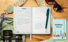 Stillleben einer Reisevorbereitung auf einer Holzoberfläche. Im Zentrum liegt ein aufgeschlagenes hellblaues „Reisetagebuch Städtetrips“, in dem handschriftliche Notizen zu einem Trip nach Barcelona im März 2026 zu sehen sind. Rechts daneben ist das geschlossene Buch mit einer Skyline-Illustration (u. a. Eiffelturm, Big Ben) abgebildet. Die Szene wird durch Reiseutensilien wie eine Sonnenbrille, eine Kamera, eine grüne Umhängetasche mit gemustertem Gurt und farblich passende Stifte ergänzt.