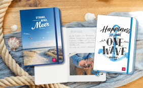 Ein stilvolles Flatlay mit Strand-Deko: Seil, Muscheln, Stoff und drei Reisetagebüchern. Links ein blaues Hardcover-Journal (GROH) mit dem Titel "STRAND, WIND, Meer" und einem Foto von Strand und Dünen. Mittig ein offenes Journal mit einem handschriftlichen Tagebucheintrag vom 15.05.2026 und einem kleinen, eingeklebten Foto von Händen, die viele kleine Muscheln halten. Rechts ein weißes Hardcover-Journal (GROH) mit einem blauen Aquarell-Anker und dem Schrifttitel "Happiness is just ONE WAVE away."