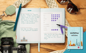 Ein Flatlay-Szene auf einem Holztisch mit Reise-Accessoires und Journalen. Ein offenes Notizbuch zeigt auf der linken Seite detaillierte handschriftliche Reiseeinträge und auf der rechten Seite ein Sterne-Rating-Formular ("Wetter", "Essen", "Unterkunft" etc.) mit lilafarbenen Stiftmarkierungen. Um das Notizbuch herum liegen persönliche Gegenstände wie eine analoge Canon-Filmkamera, eine Sonnenbrille und ein grüner Rucksack mit gemusterten Trägern. Unten rechts liegt ein geschlossenes Hardcover-Journal mit d