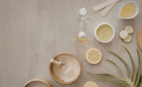 Draufsicht auf verschiedene natürliche Wellness- und Kosmetikzutaten, die auf einer grauen Steinfläche arrangiert sind. Zu sehen sind Holzschalen mit Pulver und Badesalz, eine Aloe-Vera-Pflanze, eine Zitronenscheibe, Gurkenscheiben, Öle in Fläschchen sowie Cremes und Holzspatel. Die linke Bildhälfte bietet freien Raum für Text.