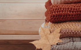 Stapel aus dick gestrickten Pullovern in Herbstfarben (Rostrot, Gelb-Rosa und Grau) auf einer Holzoberfläche, davor liegt ein trockenes, braunes Ahornblatt.