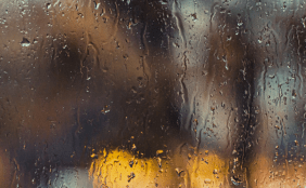 Regentropfen und Rinnen auf einer Glasscheibe mit verschwommenem, warmen Licht im unscharfen Hintergrund.
