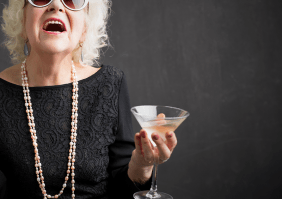Eine ältere Frau mit weißen Haaren, einem schwarzen Kleid, lange Perlenketten und Sonnenbrille hält ein Martiniglas in der Hand