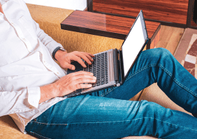 Mann in blauer Jeans und weißem Hemd sitz in einem Sessel mit einem Laptop auf dem Schoß