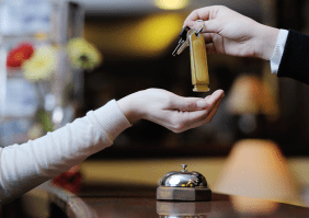 Abbildung einer Frauenhand, die einen Zimmerschlüssel von einem Mann entgegen nimmt, an einer Rezeption in einem Hotel