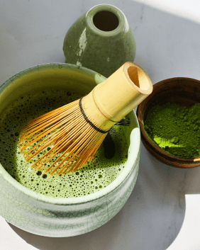 Ein Bild von der Zubereitung von Matcha-Tee, links eine Schale mit Besen und aufgeschlagenem Matcha, rechts das Pulver