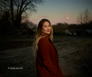 Porträtfoto von Miriam Georg, aufgenommen von Gaby Gerster. Sie steht im Freien bei Dämmerung, trägt einen rostbraunen Mantel und lächelt über die Schulter.
