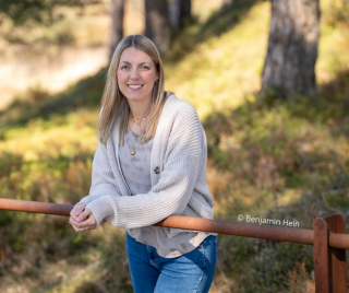Tine Nell lächelt freundlich in die Kamera, während sie sich im Freien auf ein rostbraunes Geländer stützt. Sie trägt eine beige Strickjacke über einem grauen Oberteil und Jeans. © Benjamin Hein