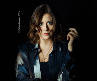 Frontales Porträt von Emily Rudolf, die mit ernstem Blick und blauen Augen direkt in die Kamera schaut, vor einem schwarzen Hintergrund. © Gaby Gerster 2023