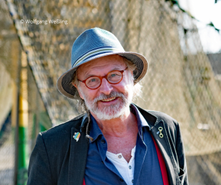 Porträtaufnahme des Autors Klaus-Peter Wolf, der freundlich in die Kamera lächelt. Er trägt einen blauen Hut, eine rötliche Brille, einen grauen Bart sowie ein dunkles Sakko, unter dem ein blaues Hemd und rote Hosenträger zu sehen sind. Oben links befindet sich der Copyright-Hinweis „© Wolfgang Weßling“.