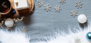 Ein gemütlicher, winterlicher Hintergrund in der Draufsicht auf einer graublauen Textiloberfläche, die viel freien Platz für Text bietet. Am oberen linken Rand steht ein Holztablett mit einer Tasse Tee und einem kleinen, weißen Deko-Häuschen. Der restliche Bereich ist weihnachtlich dekoriert mit silbernen Deko-Schneeflocken, weißen Glitzer-Weihnachtskugeln und einem kuscheligen, weißen Kunstfell-Besatz am unteren Bildrand.