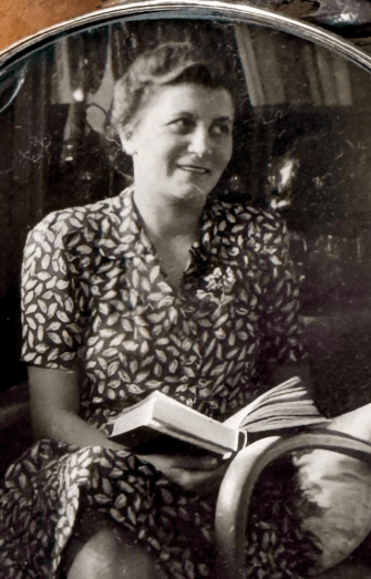 Schwarzweißfoto von Hedy. Sie lächelt und hält ein Buch im Schoß, während sie in einem Sessel sitzt. Blattgemustertes Kleid.