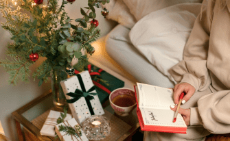 Frau sitzt im Anschnitt mit einem Pullover bekleidet auf einem Bett und hält ein Buch und einen Stift in der Hand. Die Szenerie ist weihnachtlich dekoriert
