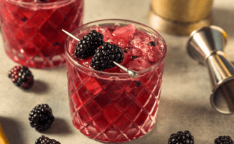 Bramble Cocktail mit Brombeeren garniert, im Hintergrund Brombeeren und Cocktail-Werkzeug