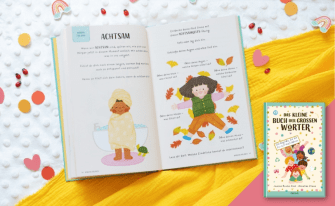 Das Bild zeigt das aufgeschlagene Buch "Das kleine Buch der großen Wörter" auf einem weißen Untergrund mit gelben Stoff. Um das Buch herum sind kleine Herzen und Regenbögen verteilt.