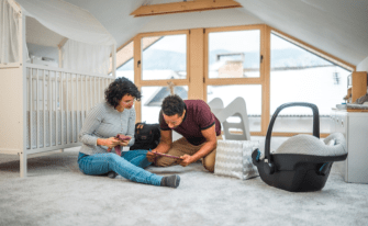 Werdene Eltern sitzen im Kinderzimmer neben dem Babybett und der Kinderschale und schreiben etwas in ein Buch