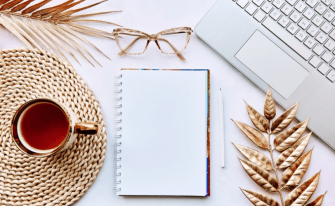 Draufsicht auf einen Schreibtisch mit Notizbuch, Stift, Laptop, Brille und goldfarbenen Dekoelementen.