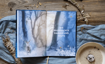 Aufgeschlagenes Buch mit Doppelseite im winterlichen Wald auf mystischen Hintergrund. Es ist Räucherwerk und eine Brennschale zu sehen auf einem blauen Tischläufer
