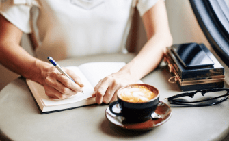 Eine Person sitzt an einem Tisch und schreibt in ein Notizbuch. Daneben stehen eine Tasse Cappuccino, eine Brille und ein Smartphone auf einer kleinen dekorativen Box.