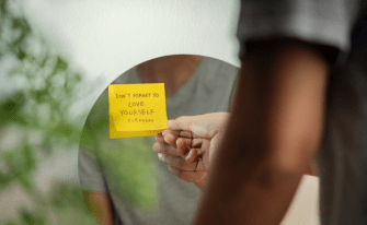 Eine Hand klebt einen gelben Notizzettel mit der Aufschrift „Don't forget to love yourself everyday“ an einen runden Spiegel, in dem sich die Person vage spiegelt.