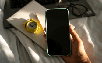 Eine Hand hält ein Smartphone mit schwarzem Bildschirm. Im Hintergrund liegen ein Notizblock, ein gelber Wecker und eine Brille auf einem hellen Laken.