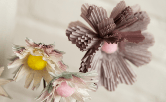 Nahaufnahme von kunstvollen, selbstgebastelten Papierblumen. Die Blütenblätter bestehen aus pastellfarbenen Papier-Muffinförmchen mit weichen Pompons als Blütenmitte in Gelb und Rosa.