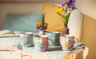 ier verschiedene Keramiktassen in Pastelltönen (Rosa und Hellblau) stehen auf einem kleinen Tisch. Die Tassen sind mit Goldrand, Blumenmotiven, Schmetterlingen und Texten wie „Kleine Auszeit“ oder „Zeit für mich“ verziert.