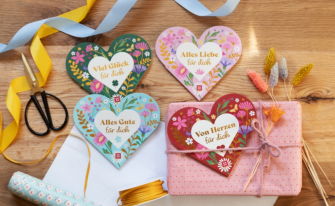 Vier herzförmige Geschenkbücher oder Karten sind dekorativ auf einem Holztisch arrangiert. Jedes Herz hat ein individuelles florales Design in frühlingshaften Farben und trägt einen positiven Schriftzug: „Viel Glück für dich“ (grün), „Alles Liebe für dich“ (hellrosa), „Alles Gute für dich“ (hellblau) und „Von Herzen für dich“ (dunkelrot). Die Szene wird ergänzt durch eine Schere, gelbe und blaue Dekobänder, ein in rosa Papier eingewickeltes Geschenk sowie getrocknete Blumen.