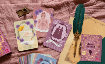 Romantisch gestaltete Orakelkarten im Regency-Stil auf einem altrosafarbenen Leinentuch. Zu sehen sind filigrane Illustrationen, eine Silhouette und eine edle, grüne Schreibfeder mit goldener Halterung neben der violetten Box „Das Jane Austen Orakel“.