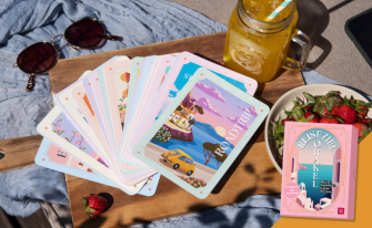 Sommerliche Szene auf einer Picknickdecke mit einer fächerförmig ausgebreiteten Kartensammlung, deren oberste Karte ein buntes „Roadtrip“-Motiv zeigt. Daneben stehen eine Schale mit frischen Erdbeeren, ein Kaltgetränk und die rosa Box „Reiseziel Orakel“.