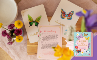 Eine helle, frühlingshafte Dekoration mit frischen Blumen und Orakelkarten, die Schmetterlinge sowie Begriffe wie „Flexibilität“, „Kreativität“ und „Verwandlung“ zeigen. Unten rechts ist die hellblaue Produktbox „Zauber der Schmetterlinge“ platziert.