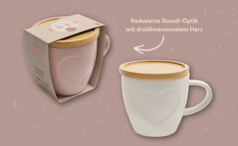Produktpräsentation des „Herzens-Bechers“ im reduzierten Skandi-Look. Links steht die rosa Tasse in ihrer Kartonverpackung, rechts daneben ist die weiße Variante unverpackt abgebildet. Beide Keramikbecher besitzen ein großes, dreidimensional eingeprägtes Herz auf der Vorderseite und sind mit einem flachen Deckel aus hellem Bambusholz verschlossen.