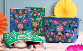 Bestickte Notizbücher in grün und königsblau mit floralem Muster, dazu passend zwei Accessoirestaschen in den gleichen Farben und Designs. Zusammen fotografiert in einem farbenfrohen Ambiente mit bunten Papierrosetten dekoriert