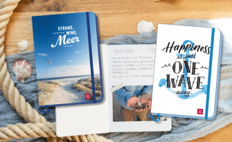 Ein stilvolles Flatlay mit Strand-Deko: Seil, Muscheln, Stoff und drei Reisetagebüchern. Links ein blaues Hardcover-Journal (GROH) mit dem Titel "STRAND, WIND, Meer" und einem Foto von Strand und Dünen. Mittig ein offenes Journal mit einem handschriftlichen Tagebucheintrag vom 15.05.2026 und einem kleinen, eingeklebten Foto von Händen, die viele kleine Muscheln halten. Rechts ein weißes Hardcover-Journal (GROH) mit einem blauen Aquarell-Anker und dem Schrifttitel "Happiness is just ONE WAVE away."